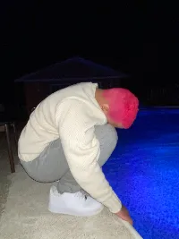 Photo of me perched up by a pool with hot pink hair with my head bowed into my knees